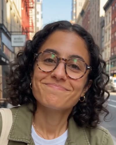 Her: Late twenties, curly shoulder-length black hair, round tortoiseshell glasses, olive utility jacket over a white tee, canvas tote on her shoulder.